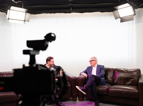 Two people sitting on a sofa in a studio environment with a camera in the foreground filming them.