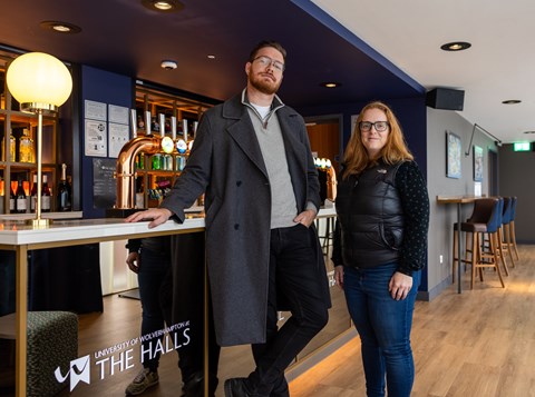 Two people stand in a modern bar area with the sign "The Halls, University of Wolverhampton" visible on the counter. Bottles and taps are displayed in the background.