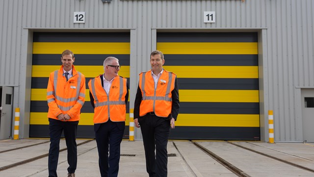 Three men, in orange hi-vis vests walking in front of large metal warehouse building which fills rest of the picture. There are two black and yellow striped garage doors and a sign above says West Midlands Railway