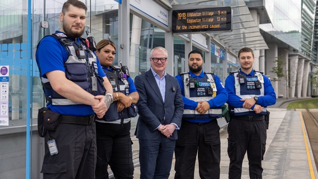 A group of five people stood under a tram stop canopy with a city centre office behind in the distance. Four of the line up are wearing Transport Safety uniforms, including bright blue polo shirts with a dark stab-proof vest over the top. In the centre of the line is Mayor Richard Parker wearing his suit jacket.