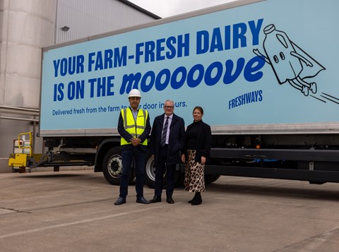 Three people standing in front of a blue lorry - the side of which bears a light blue design - with words 'your farm fresh dairy is on the moooove'. Behind that is a factory building and to the side part of steel piping and silo